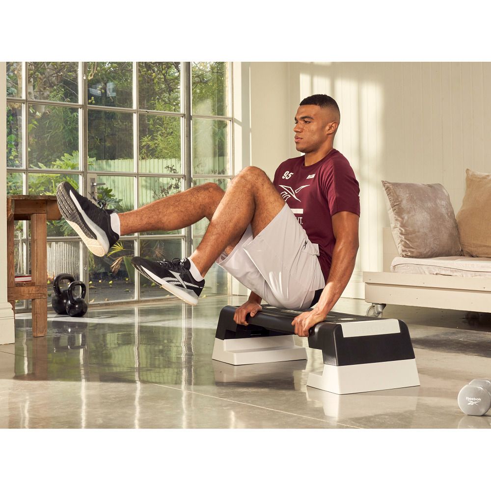 Schwarzer Reebok Step mit weißen Füßen. Mann sitzt darauf und hebt die Beine. Im Hintergrund: Fenster, Sofa, Hanteln.