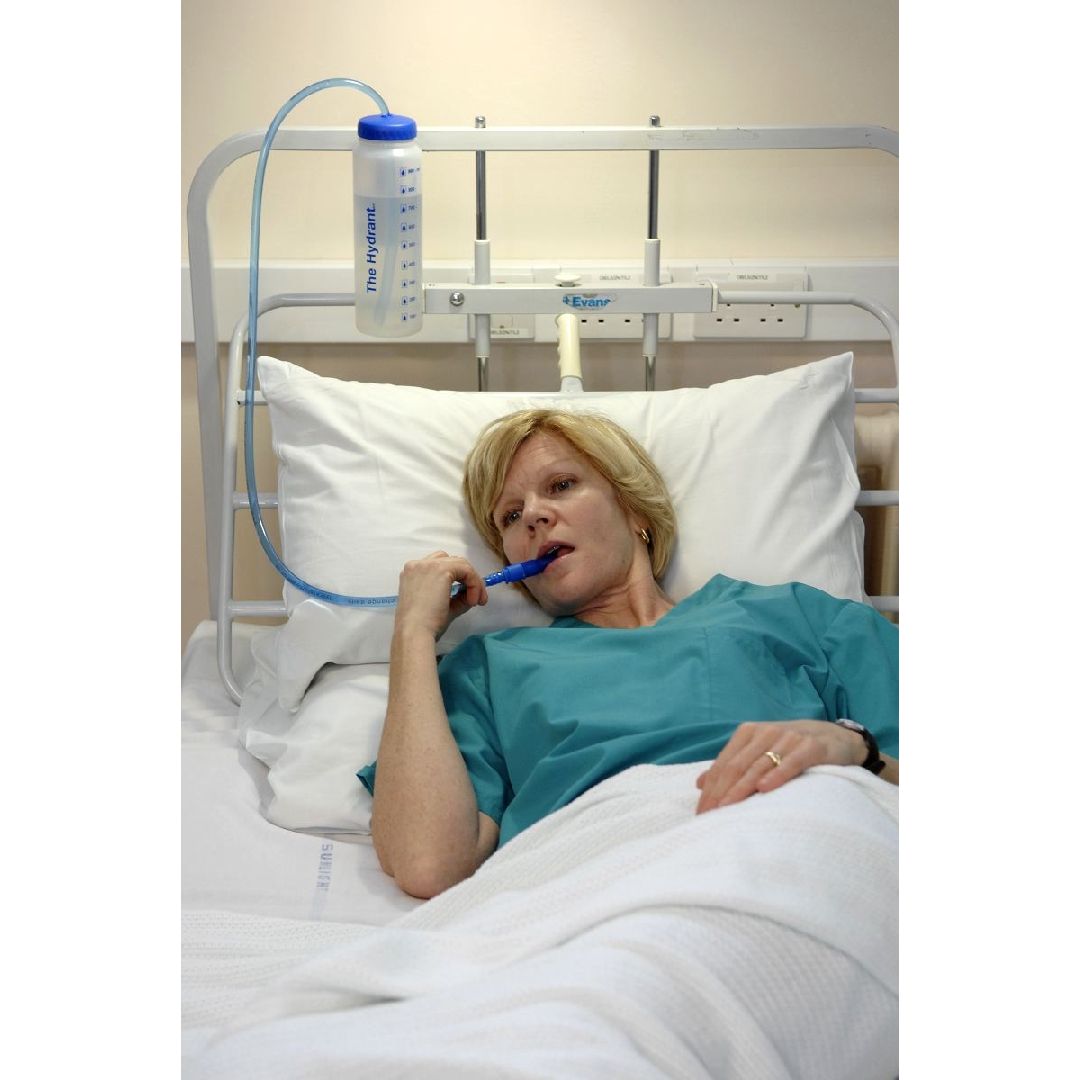 Frau im Bett, trinkt aus einer Trinkflasche mit Strohhalm. Die Flasche ist am Betthaupt befestigt.