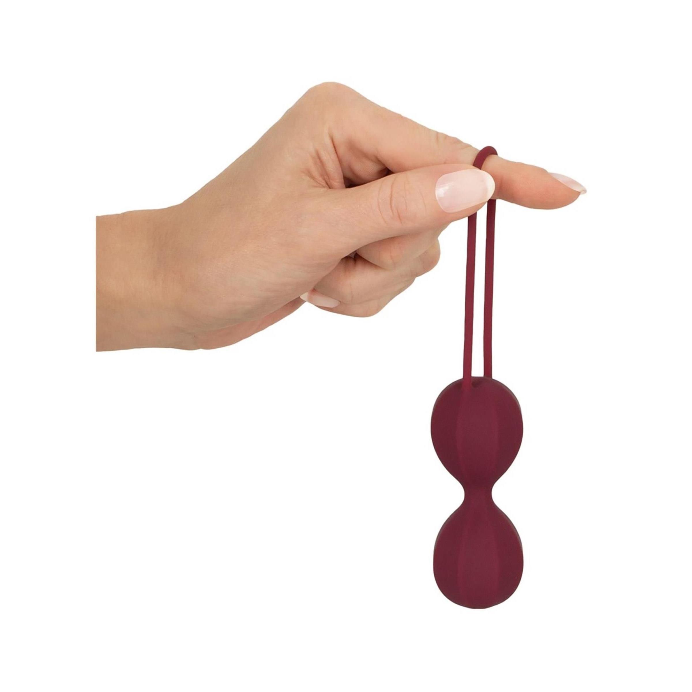 Hand hält burgunderfarbene Liebeskugel an der Schlaufe. Zwei Kugeln, gerillte Oberfläche. Weißer Hintergrund.