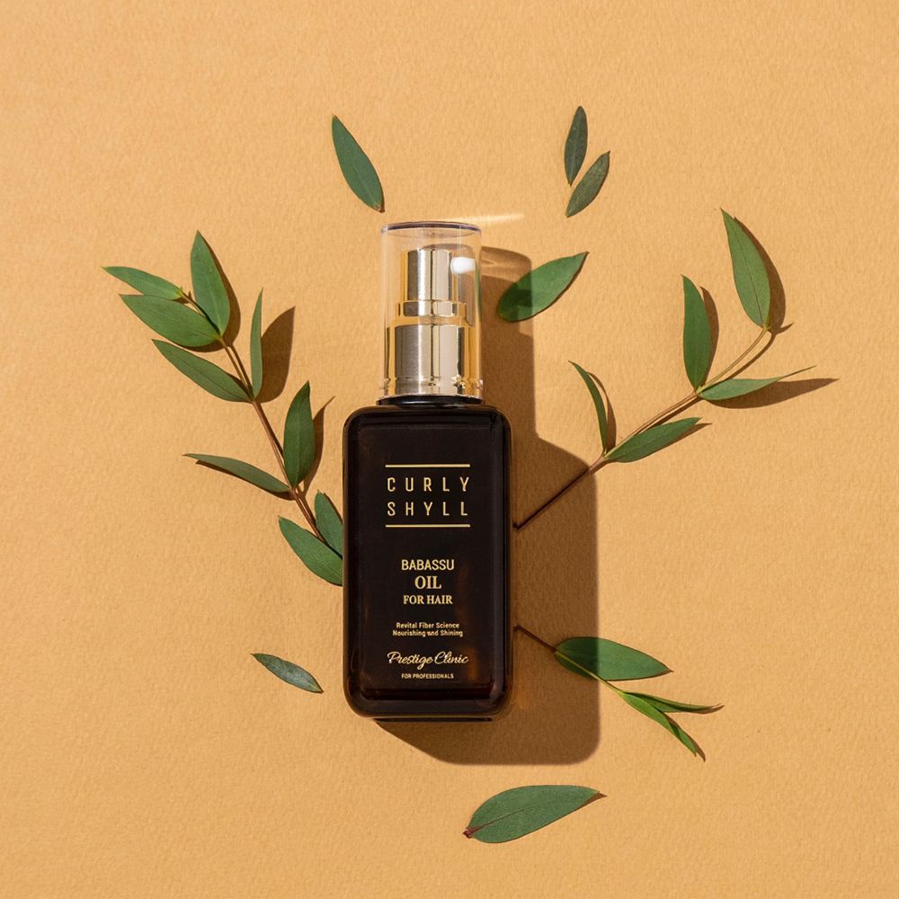 Braune Glasflasche mit goldfarbenem Sprühkopf. Aufschrift: CURLY SHYLL Babassu Oil for Hair. Umgeben von grünen Blättern.