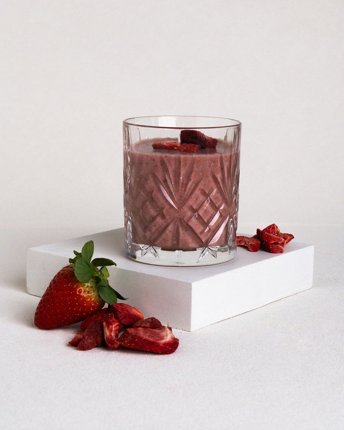 Glas mit Erdbeer-Smoothie, gefriergetrocknete Erdbeeren und frische Erdbeere. Auf weißem Podest.