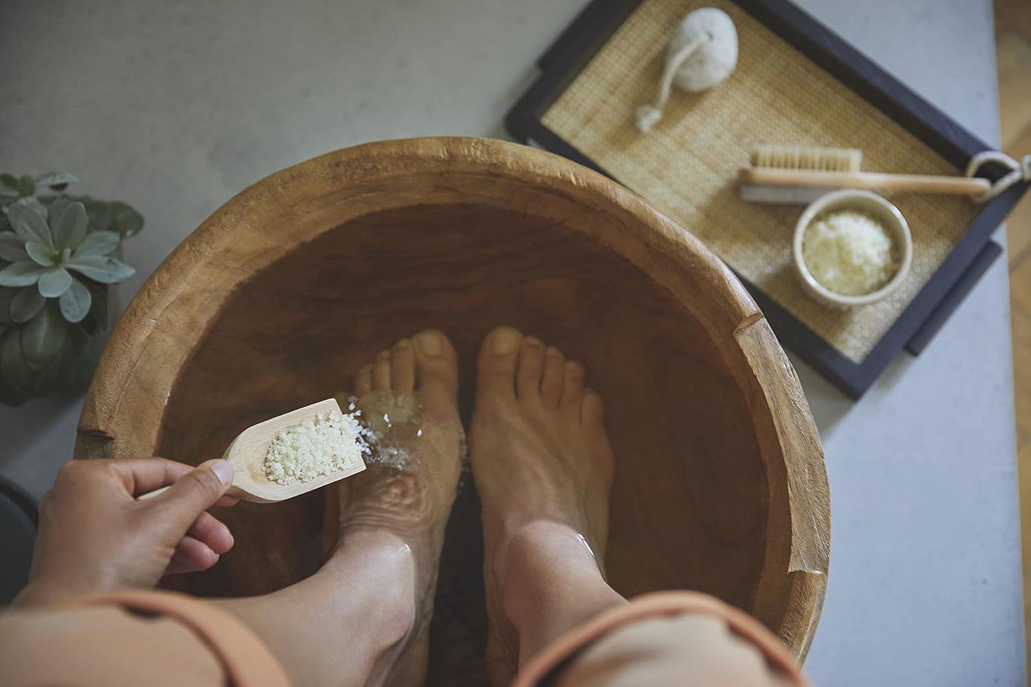 Füße in einer Holzschale mit Wasser. Person streut Kristalle mit einem Löffel auf die Füße. Zubehör: Bürste, Schwamm, Tablett.