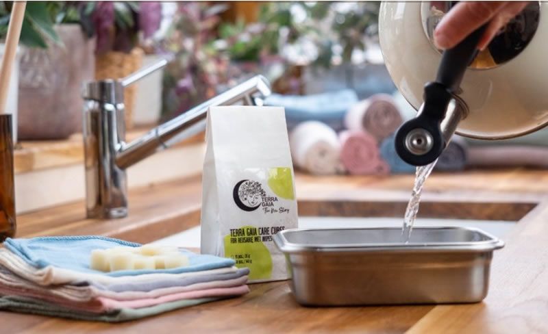 Person gießt Wasser aus einem Wasserkocher in einen Behälter. Daneben liegen Tücher, Care Cubes und eine Verpackung mit Logo.