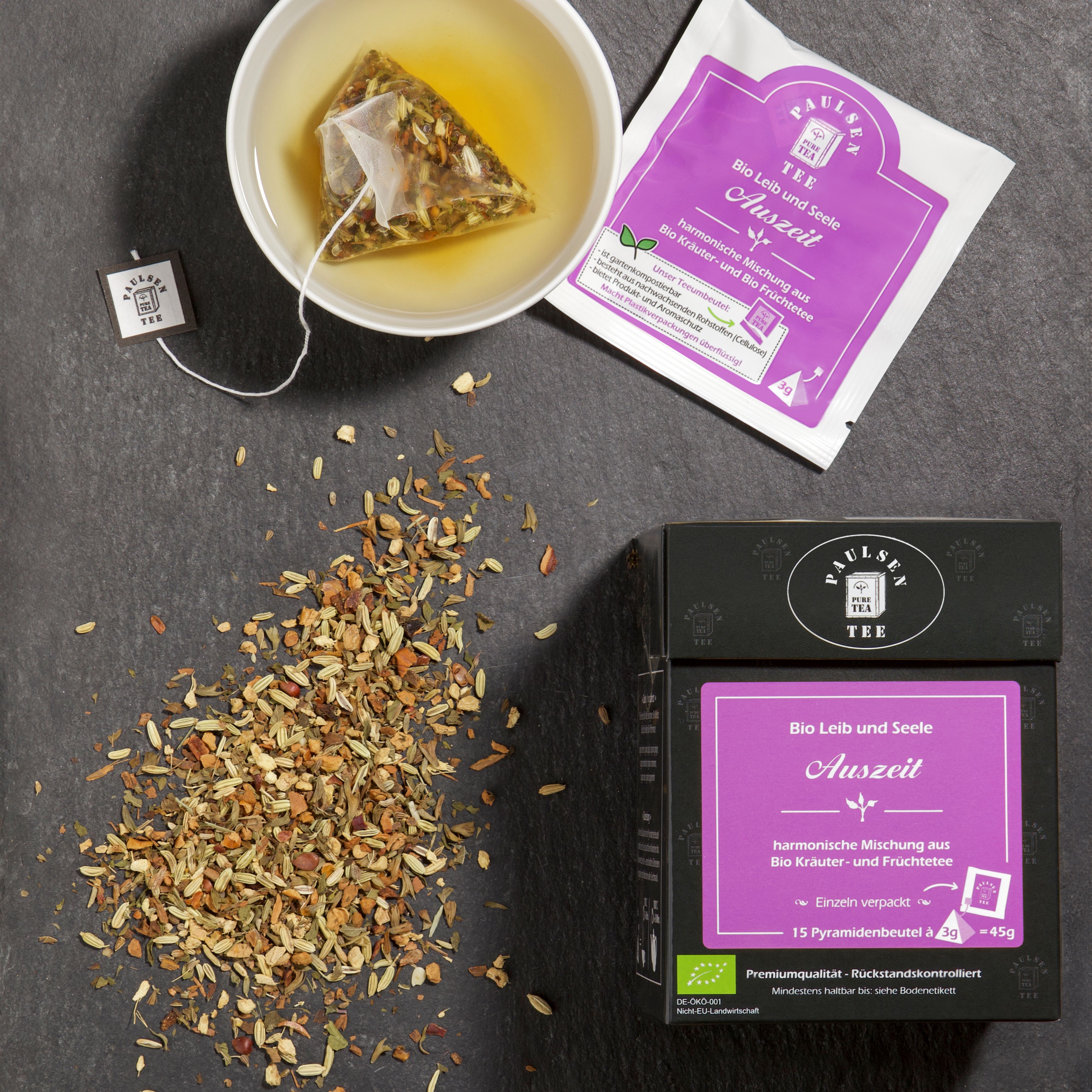 Tasse Tee mit Pyramidenbeutel. Daneben Teebeutel und Tee-Box. Tee-Box mit lila Etikett. Aufschrift: Bio Leib und Seele Auszeit.