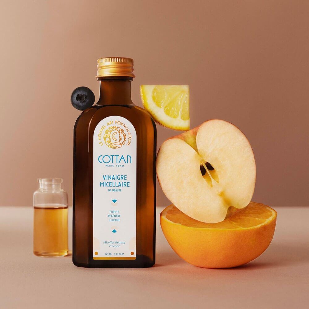 Braune Glasflasche mit goldfarbenem Verschluss. Weißes Etikett mit blauer Schrift. Neben Obststücken.