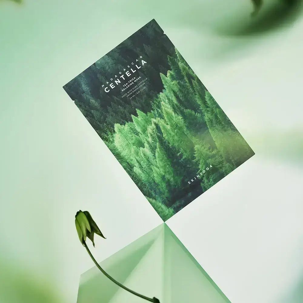 Verpackung der Gesichtsmaske mit Waldmotiv und Schriftzug MADAGASCAR CENTELLA. Liegt auf einer Glasfläche, Blume.