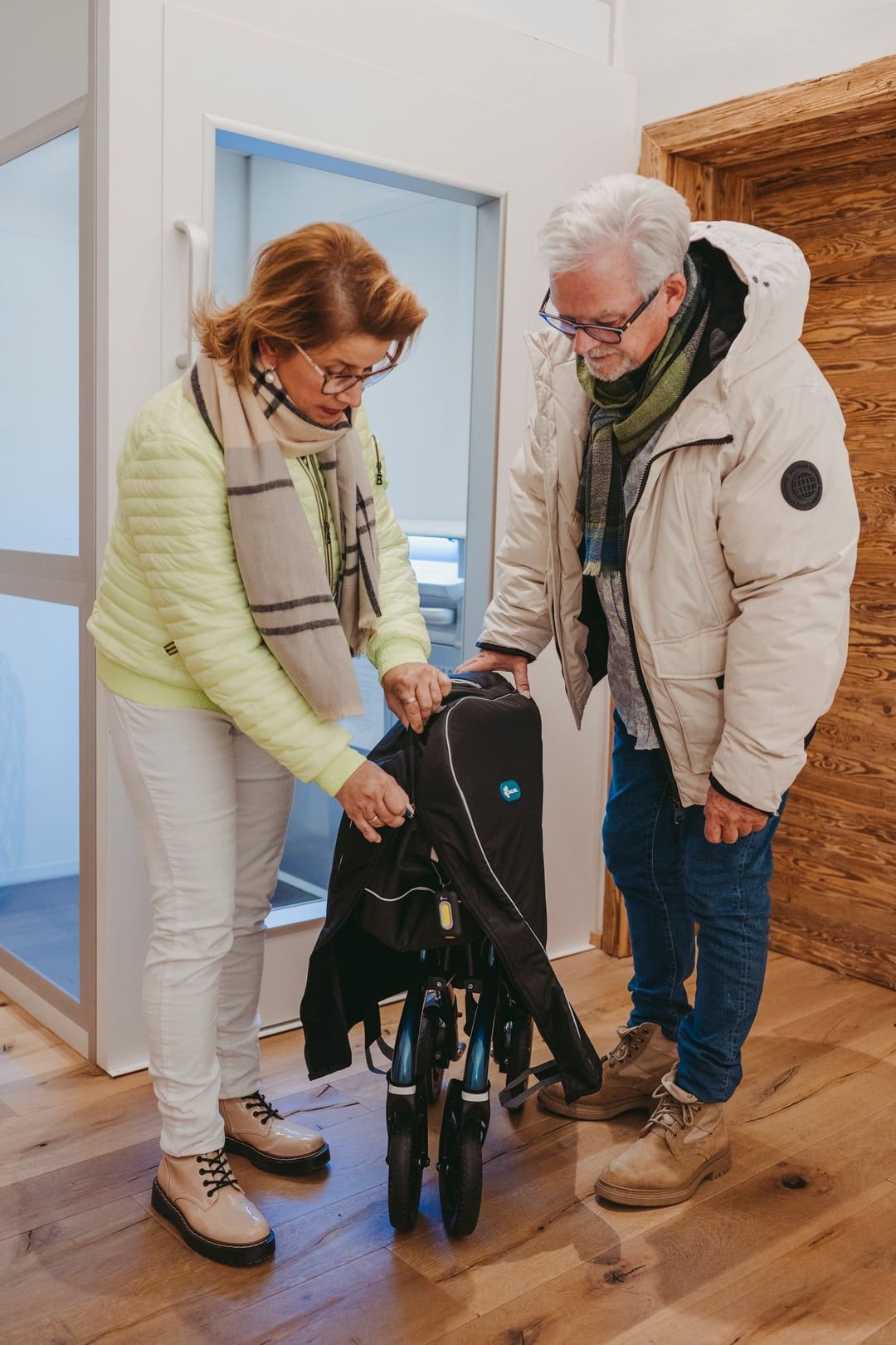 Zwei Personen falten einen Rollator zusammen. Die schwarze Tasche ist teilweise geöffnet. Holzfußboden und helle Wand.