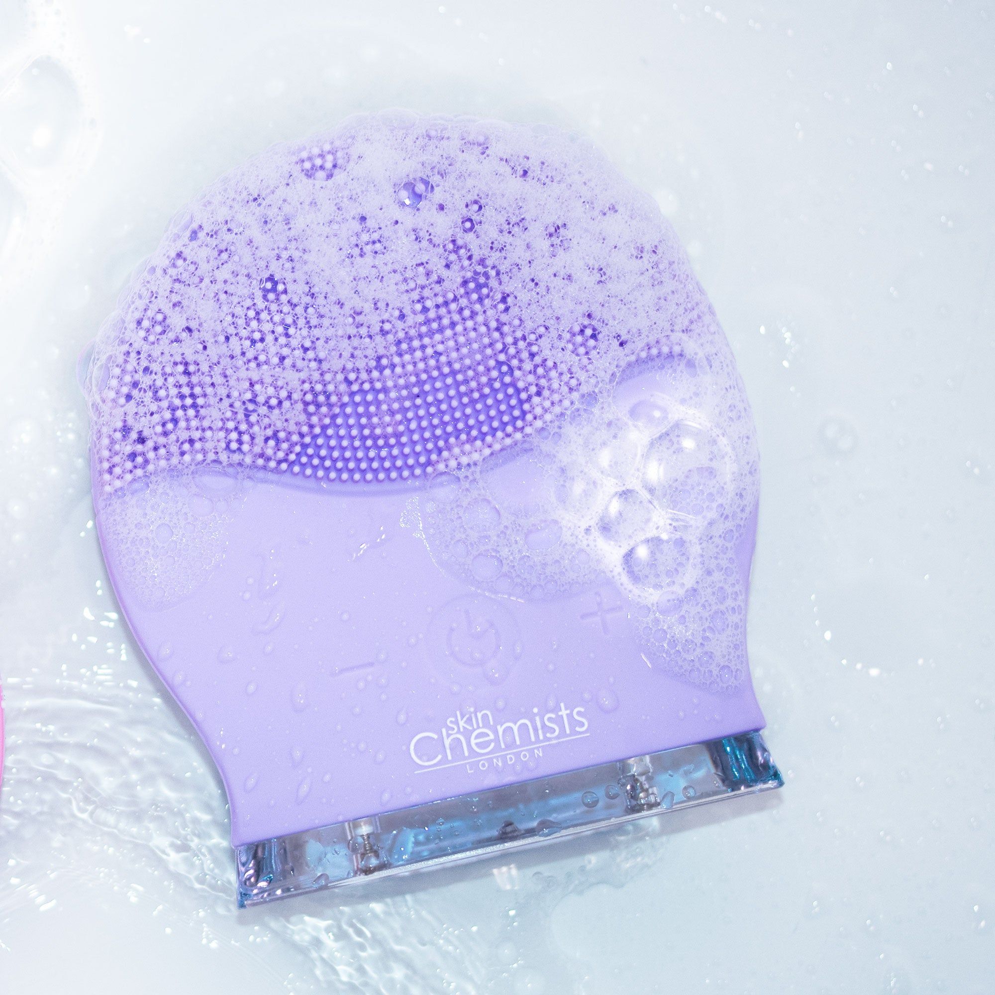 Lila Gesichtsreinigungsbürste in Wasser mit Seifenschaum. Bürste mit Silikonborsten, Logo sichtbar.
