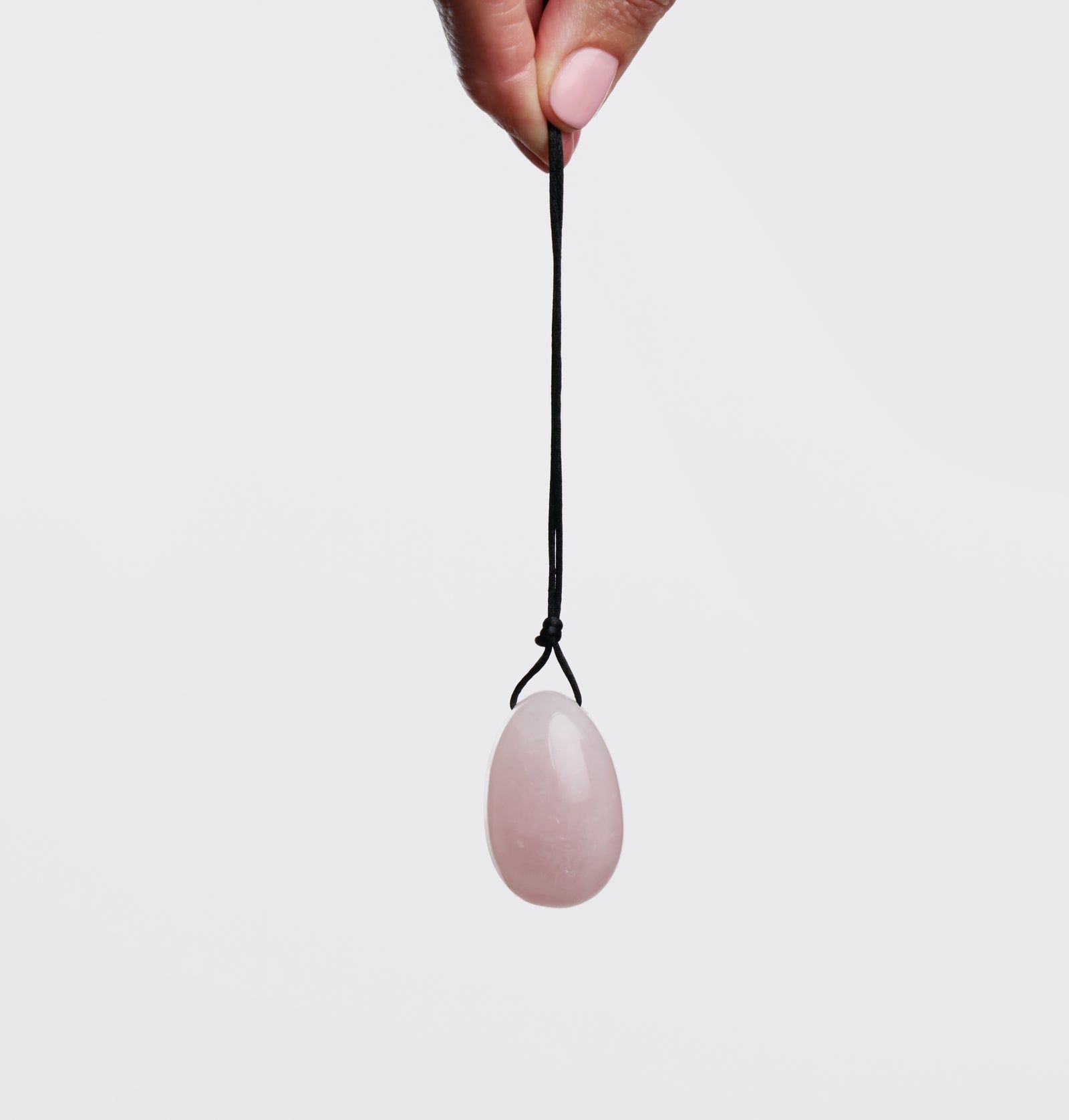 Rosafarbener Yoni-Ei aus Stein, an einer schwarzen Schnur gehalten. Die Hand einer Person hält die Schnur vor hellem Hintergrund.