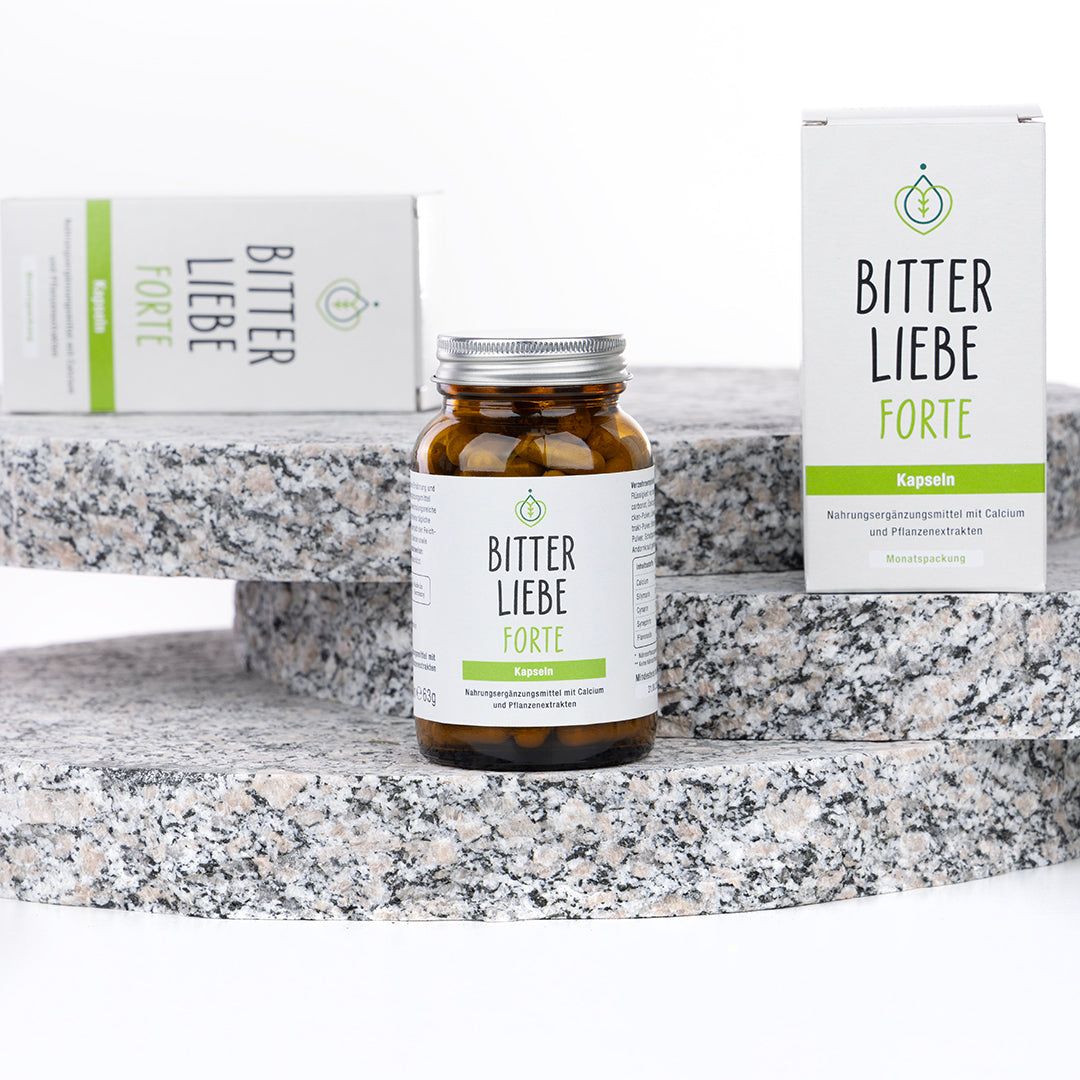 Zwei Schachteln und eine Flasche "BitterLiebe Forte" auf gestufter Oberfläche. Flasche mit silbernem Deckel.