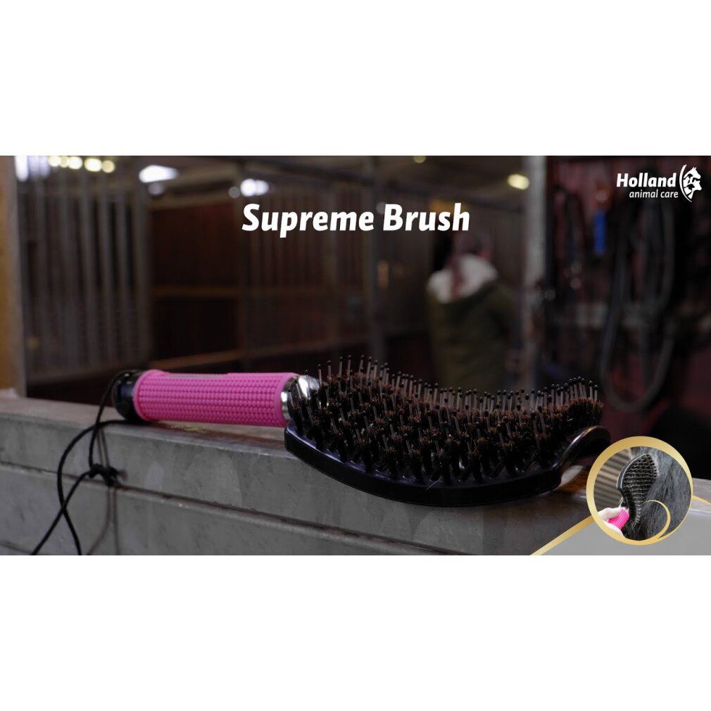 Bürste mit rosa Griff und schwarzem Bürstenkopf. Produktname: Supreme Brush. Im Hintergrund ist ein Stall zu sehen.