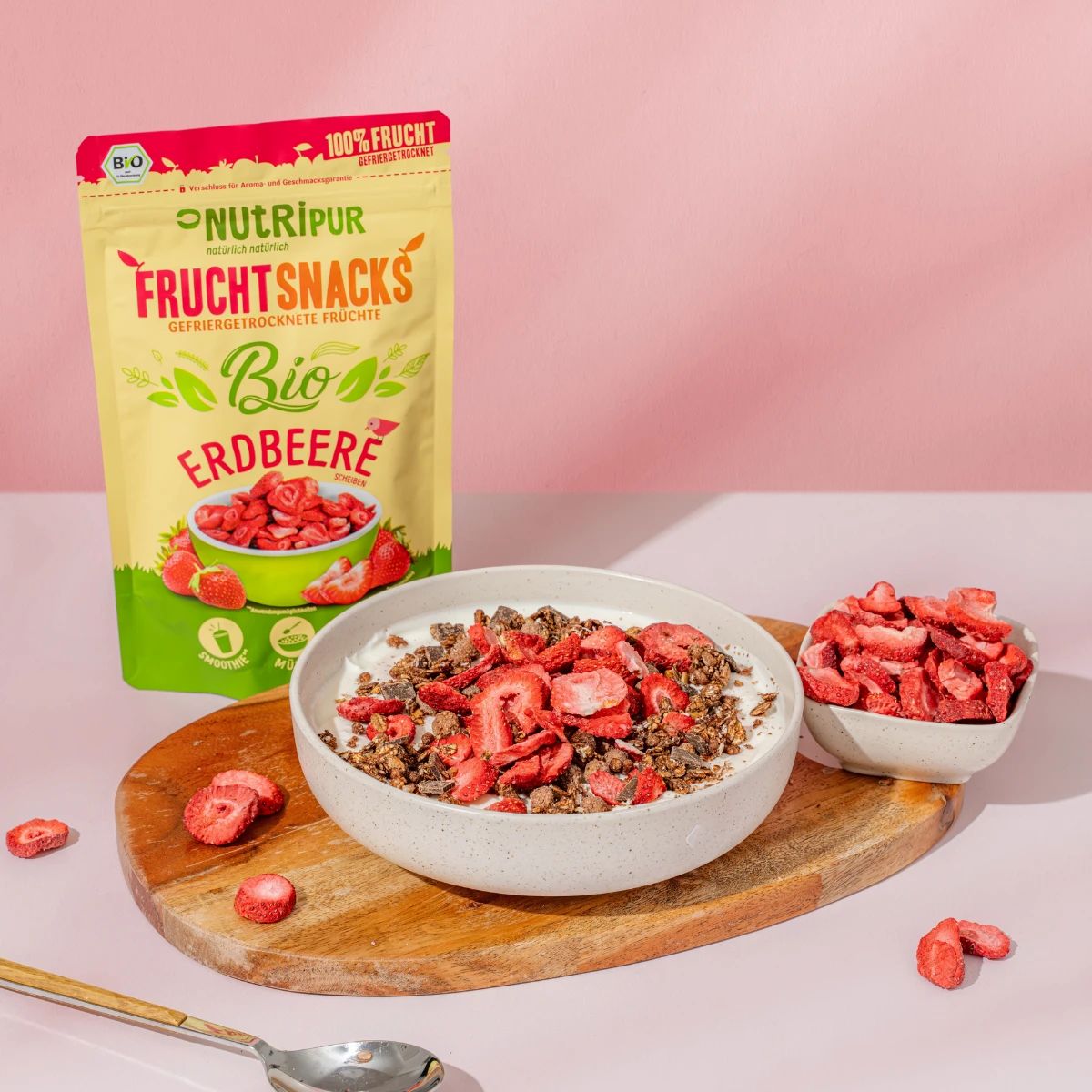 Gefriergetrocknete Erdbeeren in Schalen. Eine Schale mit Joghurt und Müsli. Im Hintergrund: Packung NutriPur Bio Erdbeeren.