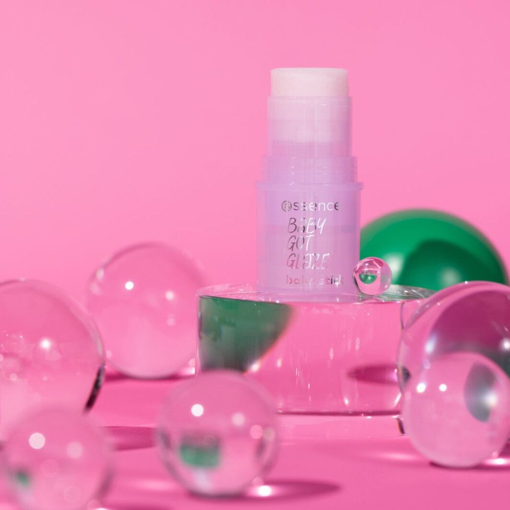 Essence Baby Got Glaze Stick Balsam auf Podest. Rosa Hintergrund mit Kugeln. Produktname und Marke sichtbar.