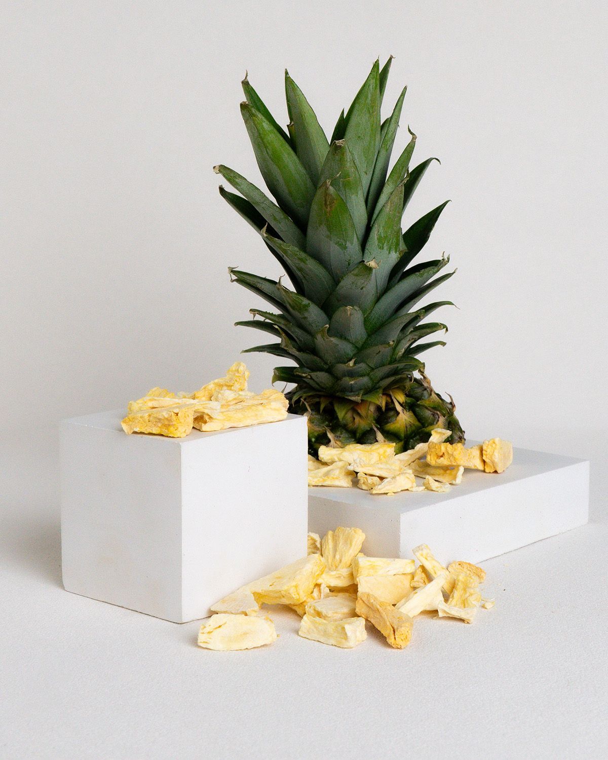 Ananas mit Blättern, neben weißen Würfeln mit gefriergetrockneten Ananasstücken. Einige Stücke liegen verstreut.