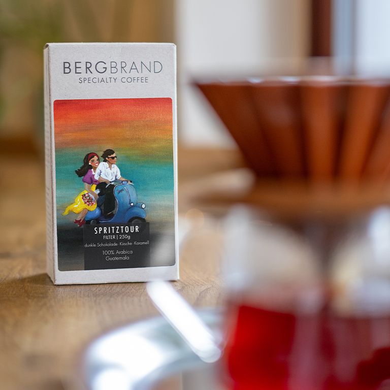 Kaffeepackung BERGBRAND Spritztour neben einer Kaffeemaschine. Illustration von Personen auf einem Roller.