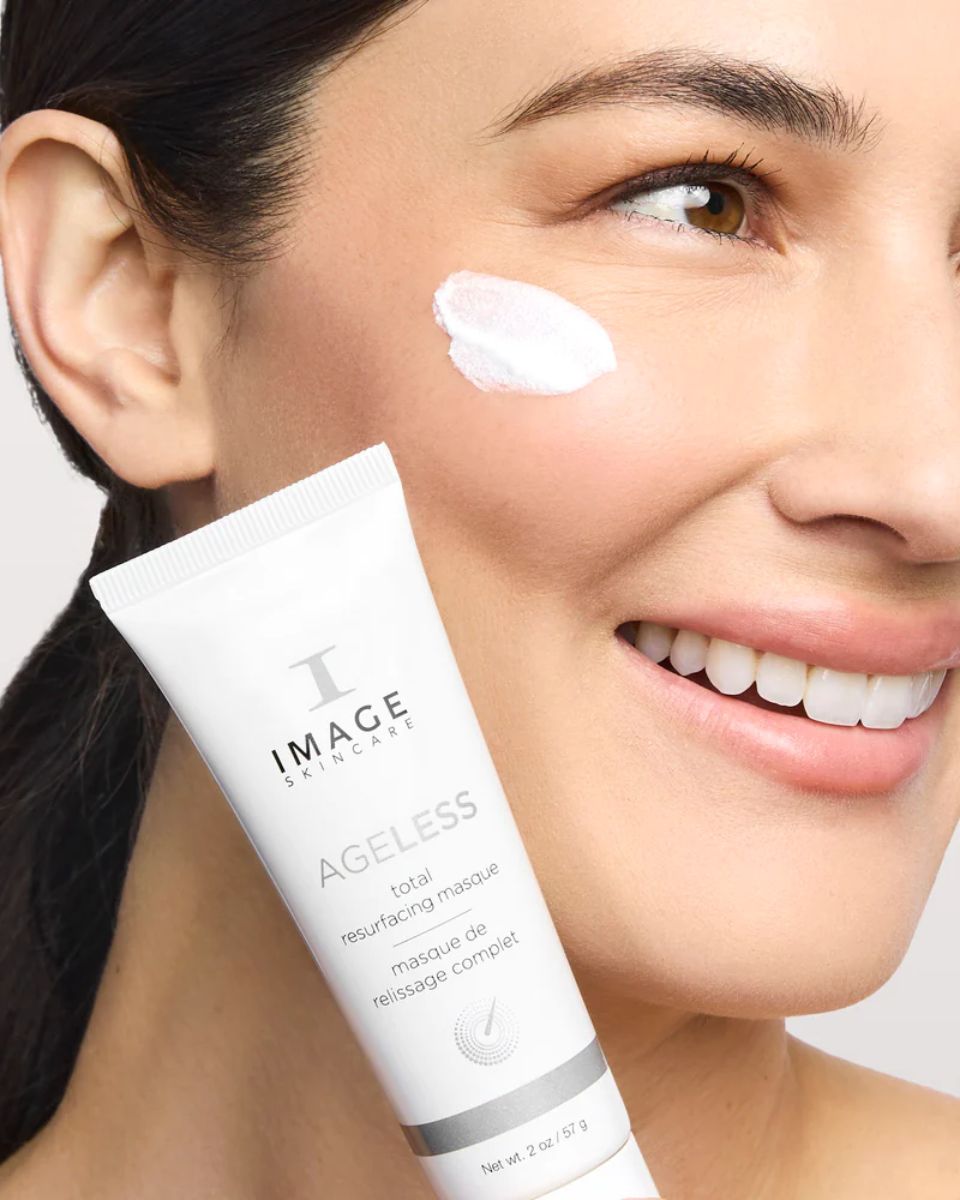 Frau mit Produkt auf der Wange. Tube: IMAGE Skincare, AGELESS total resurfacing masque.
