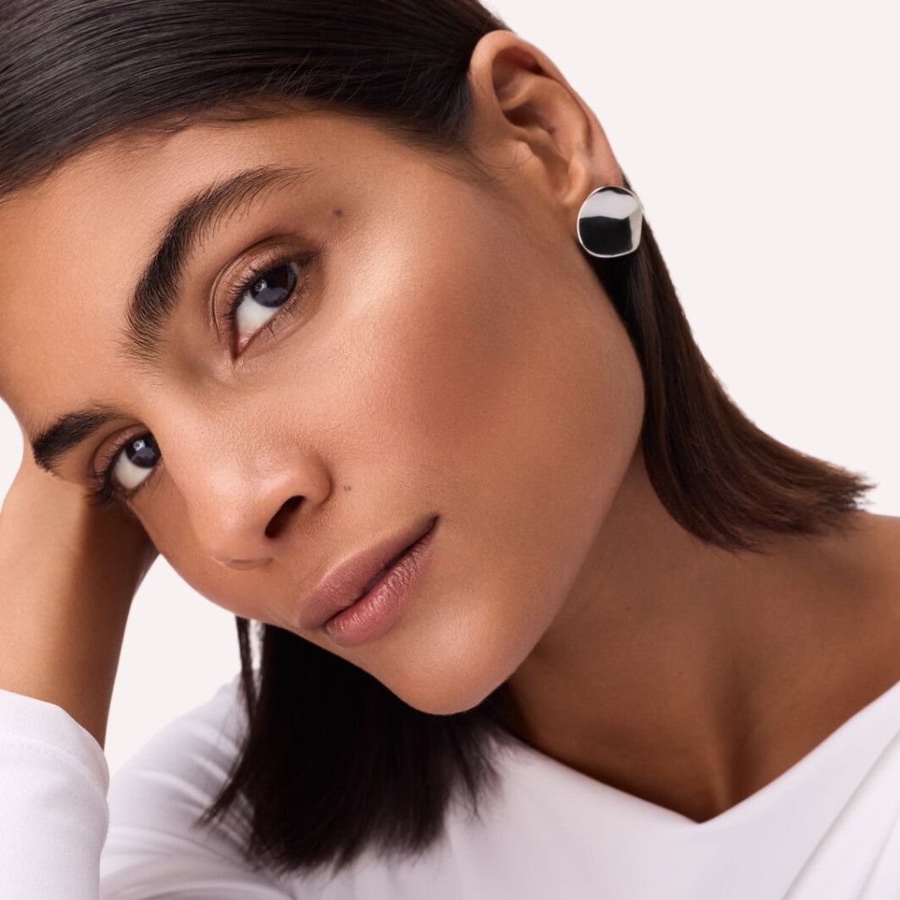 Porträt einer Frau mit dunklen Haaren. Highlighter auf Wangenknochen. Schwarze Ohrringe. Weißes Oberteil.