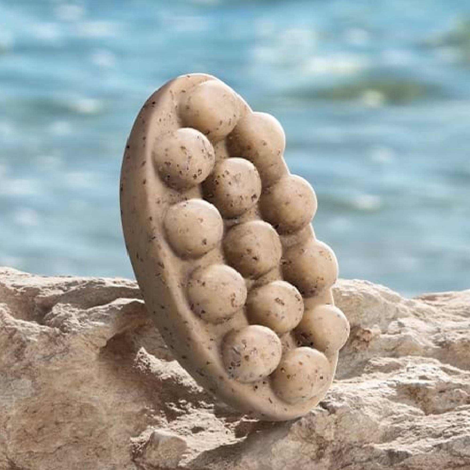 Ovale Seife mit Noppen, beige mit braunen Punkten, vor Felsen und Meer.