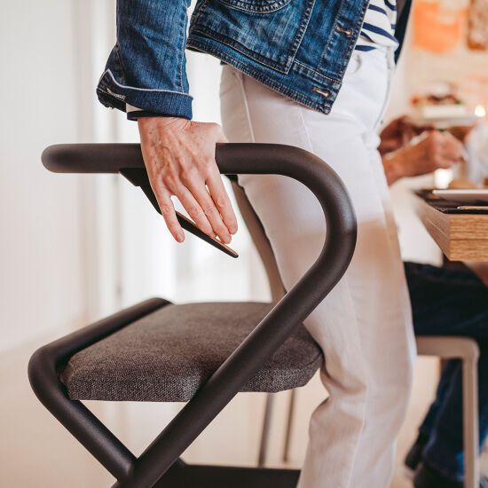 Person hält Armlehne eines grauen Rollators. Graue Sitzfläche, schwarzer Rahmen. Person trägt Jeansjacke und gestreiftes Shirt.