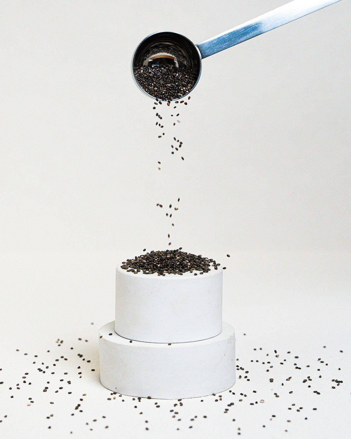 Chia Samen fallen aus einem Löffel auf eine weiße, zylindrische Oberfläche. Mehr Samen liegen verstreut.