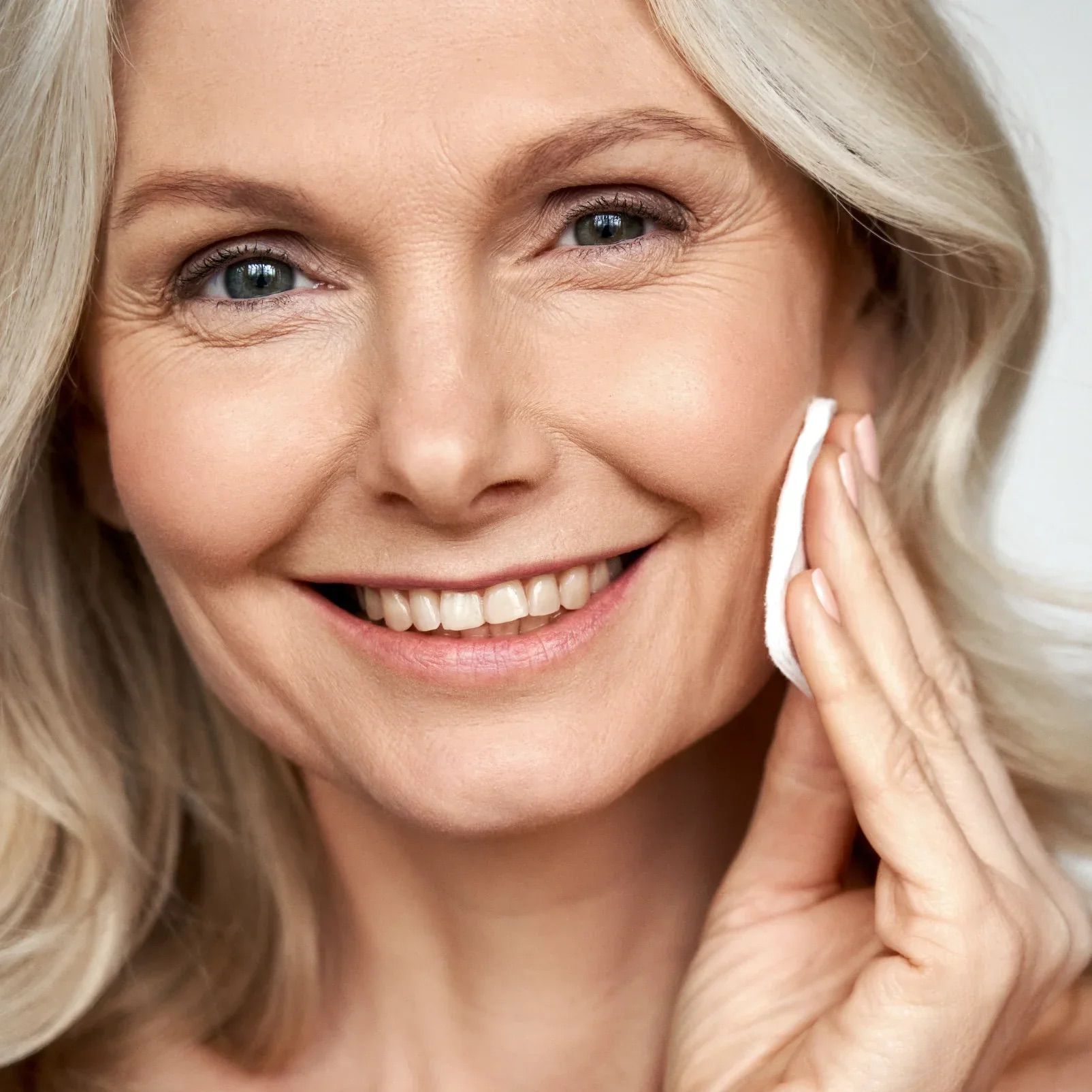 Ältere Frau mit blonden Haaren. Sie reinigt ihr Gesicht mit einem Wattepad.