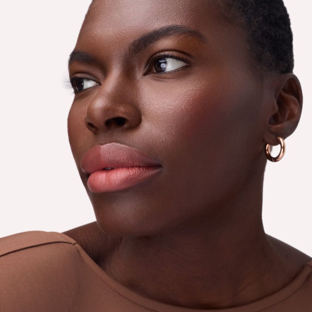 Porträt einer Frau mit rosafarbenen Lippen. Sie trägt goldene Creolen und einen braunen Pullover.