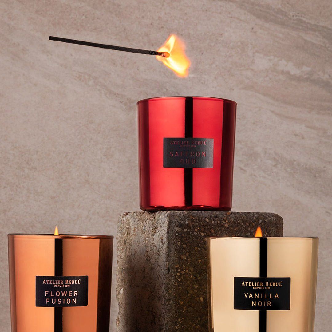Drei Duftkerzen in verschiedenen Farben. Eine brennt, eine wird angezündet. Aufschrift: Atelier Rebul, Saffron Oud, Flower Fusion, Vanilla Noir.