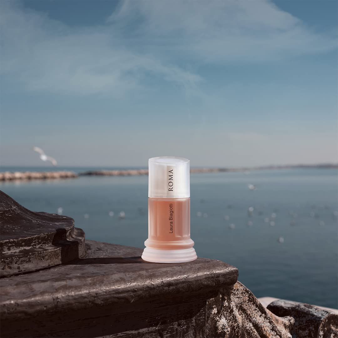 Laura Biagiotti Roma Eau de Toilette Spray. Flakon auf Steinmauer, Meer im Hintergrund. Flakon in Säulenform, hellorange.