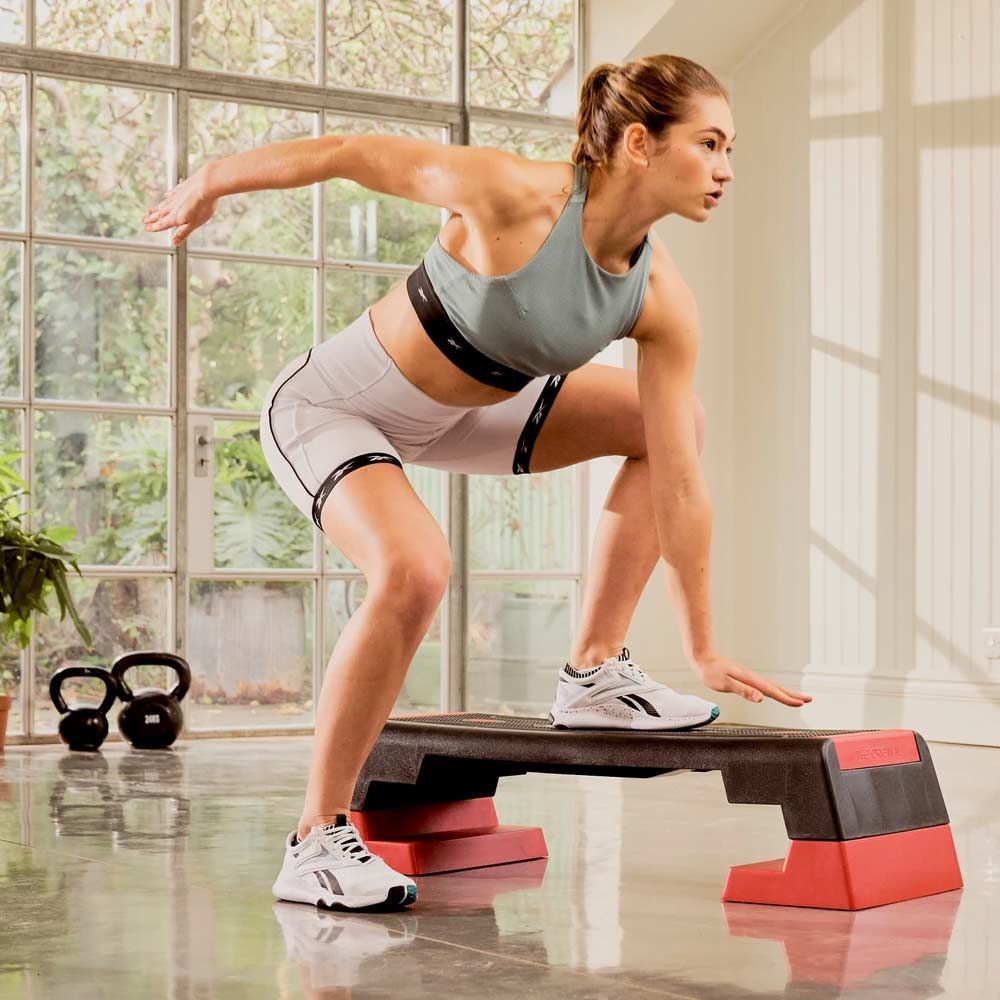 Frau trainiert auf einem Reebok Step. Sie trägt Sportkleidung. Im Hintergrund: Fenster, Kettlebells.