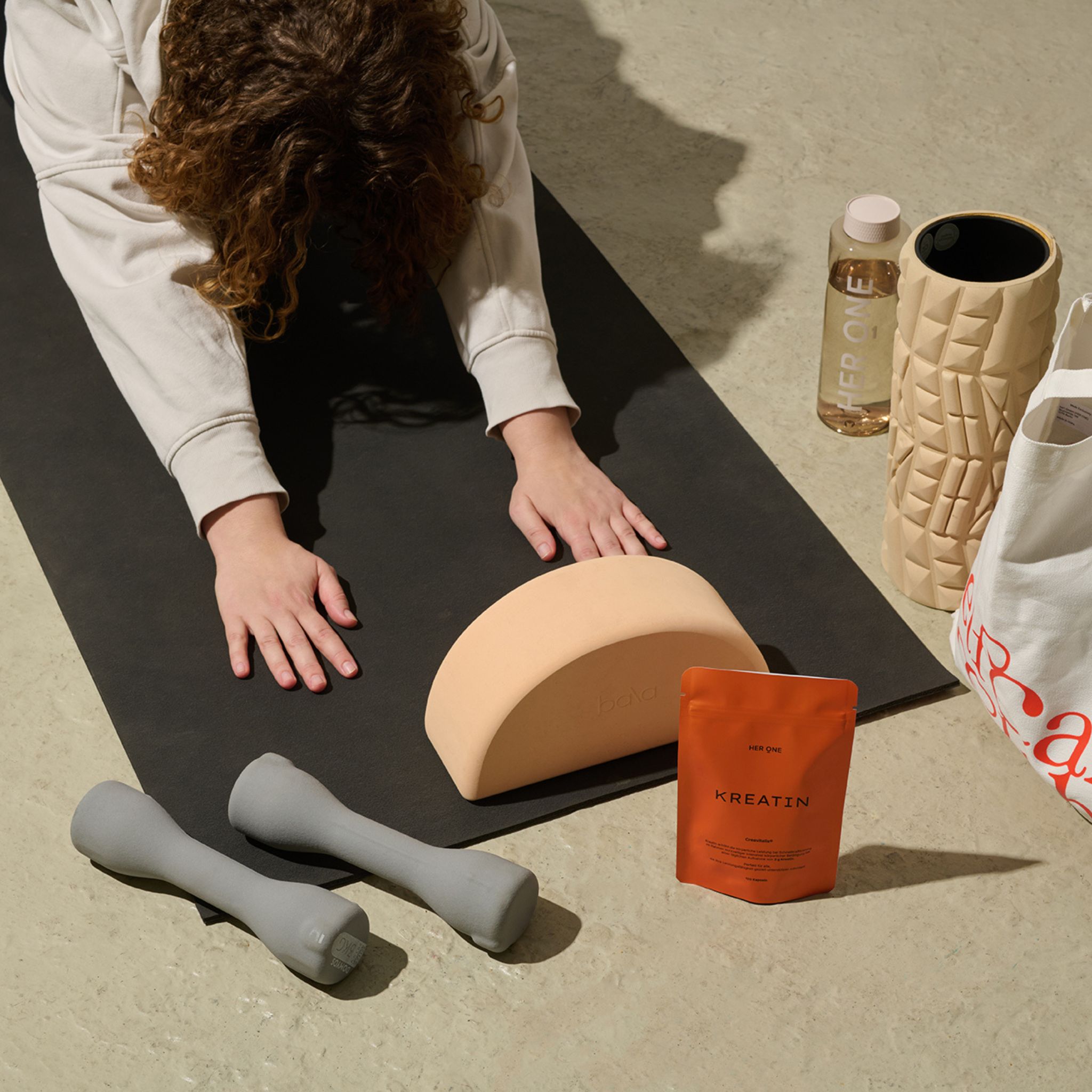 Person liegt auf einer Yogamatte. Neben ihr: "HER ONE KREATIN" Verpackung, Hanteln, Wasserflasche, Rolle.