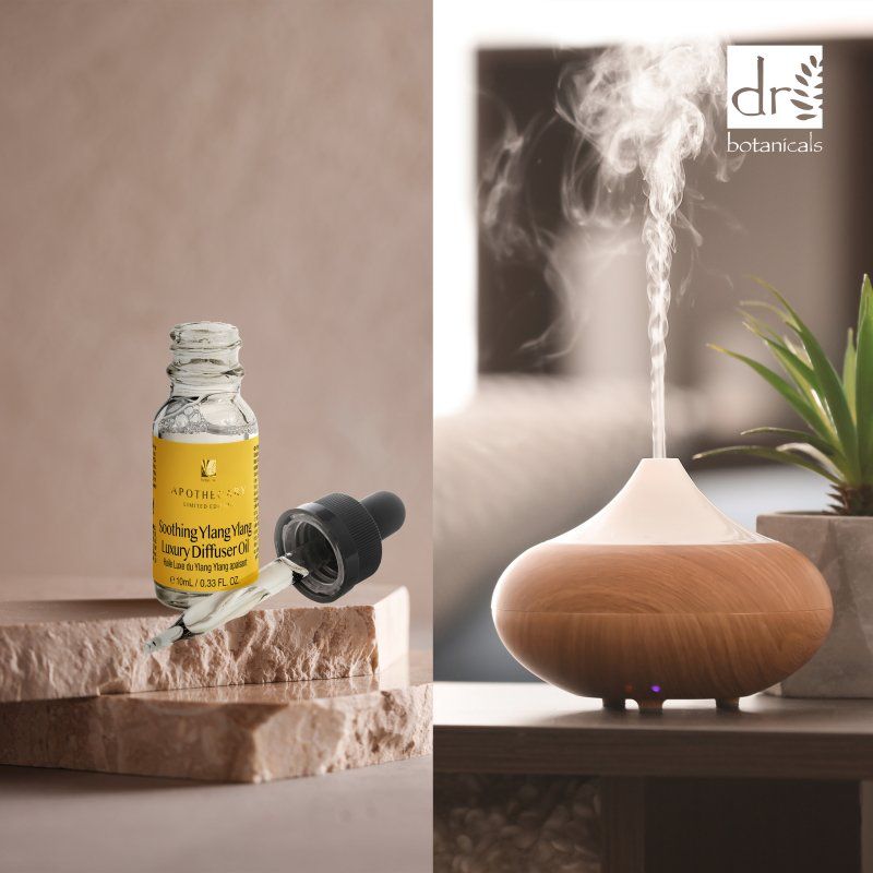 Diffusor aus Holz und weißem Kunststoff, daneben Ölflasche mit Pipette. Aufschrift: "Soothing Ylang Ylang Luxury Diffuser Oil". Dampf steigt auf.