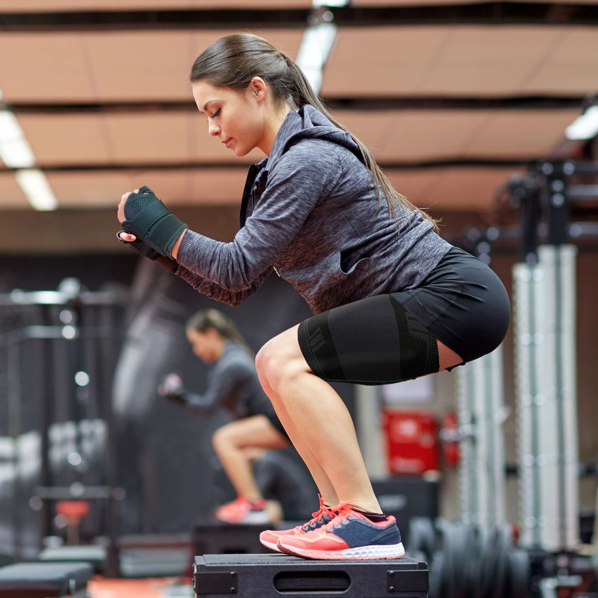 Frau beim Training mit Oberschenkelbandage. Sie macht Kniebeugen auf einer Box. Eine weitere Person im Hintergrund.