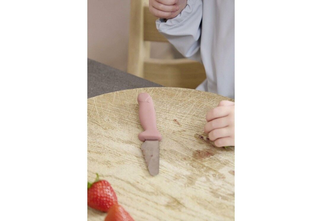 Kind schneidet auf Holzbrett mit rosa FILIBABBA Kindermesser. Erdbeere liegt daneben. Kind trägt Schürze.