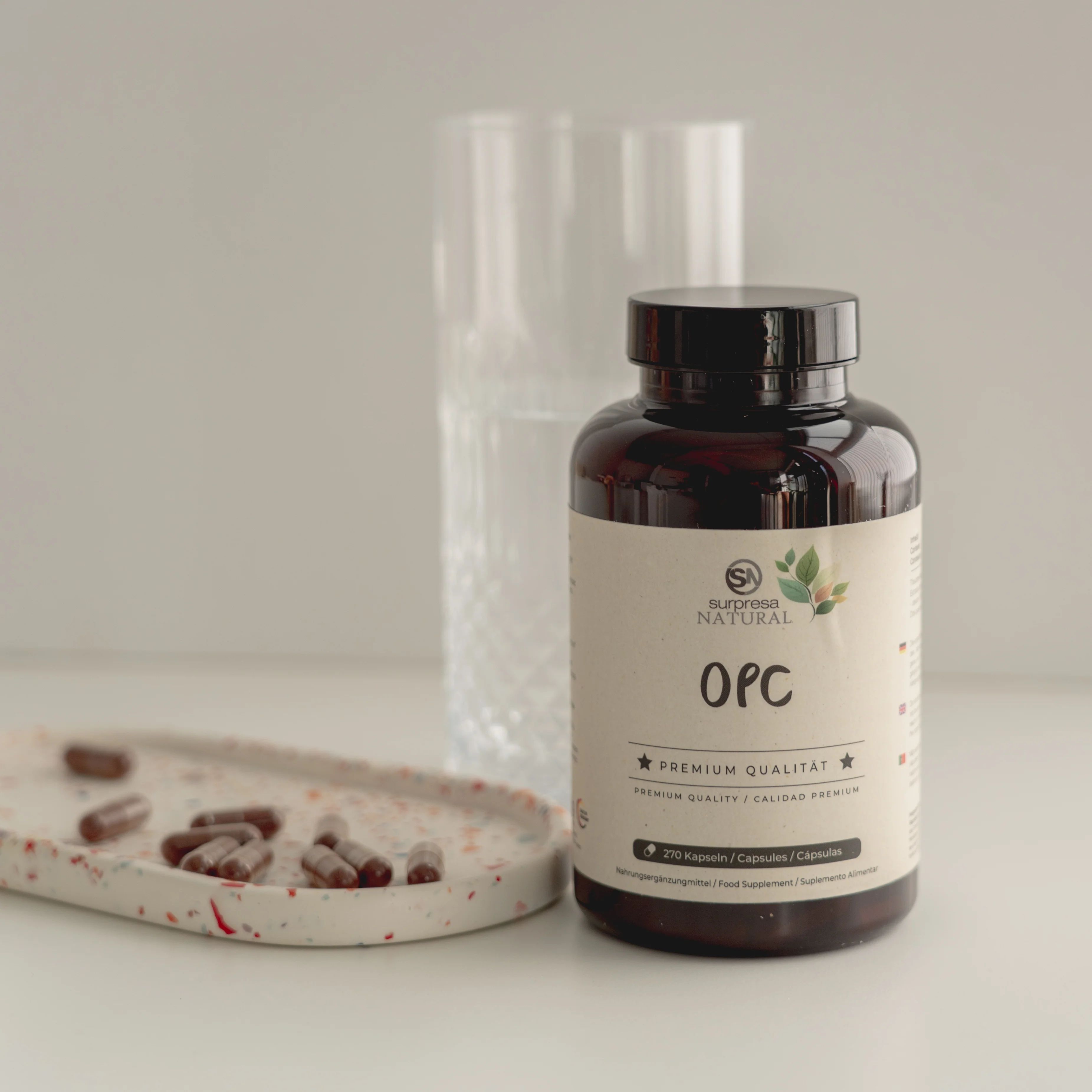 Braune Kapseln in einer Flasche und auf einem Tablett. Glas im Hintergrund. Aufschrift: OPC, Premium Qualität.