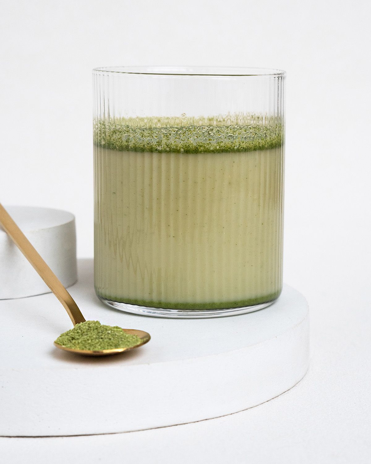 Glas mit grünem Getränk und Löffel mit Moringa-Pulver. Auf weißem Podest.