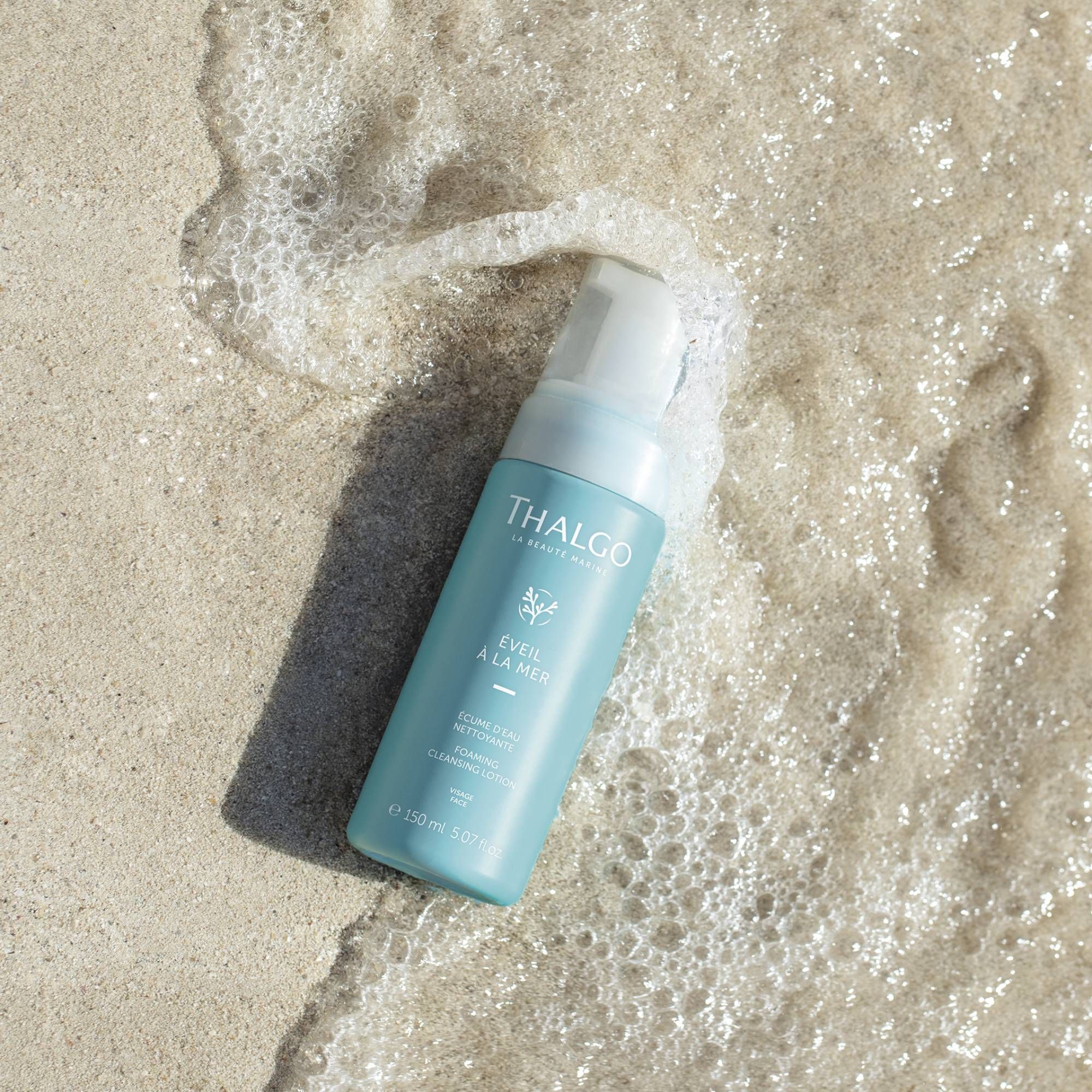 Türkisgrüne Flasche auf Sand, umgeben von Schaum. Aufschrift: THALGO, ÉVEIL À LA MER, Reinigungs-Lotion. Volumen: 150 ml.
