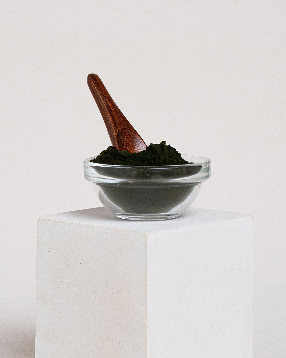 Kleine Glasschale mit grünem Chlorella Pulver. Holzlöffel steckt in der Schale. Auf weißem Podest.