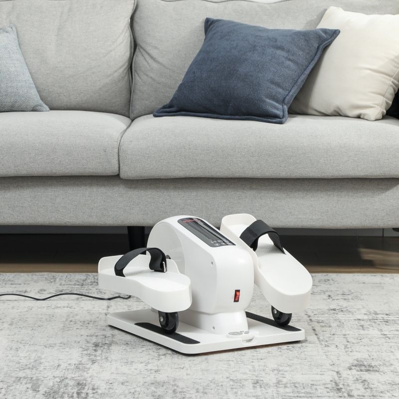 Weißes Mini-Ellipsentrainer-Fitnessgerät mit schwarzen Riemen. Auf Teppich vor Sofa. Rote Ein-/Aus-Taste, Rollen.