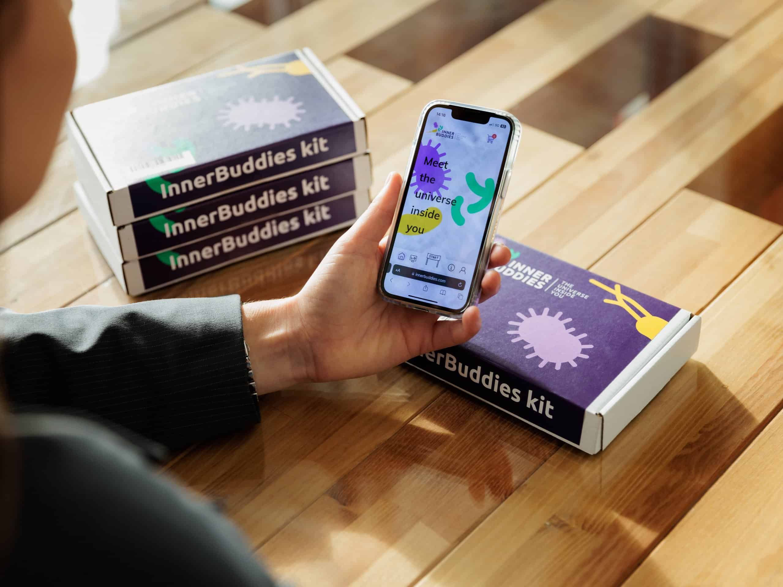 Drei gestapelte Inner Buddies Kits und ein Smartphone, das die App mit Mikroben-Grafiken zeigt.