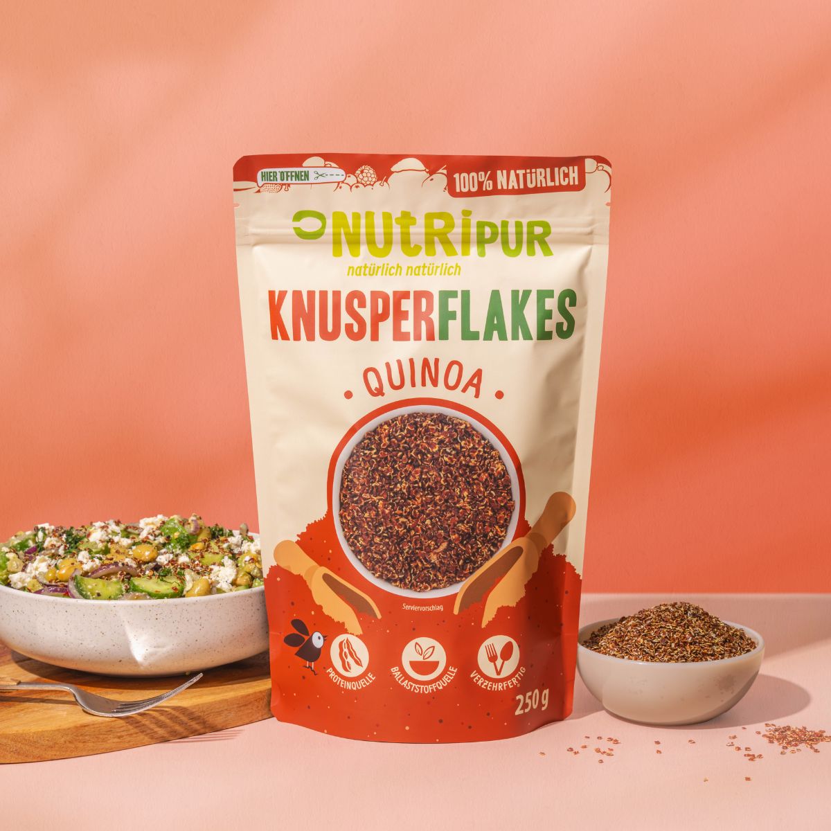 Beutel NutriPur Knusperflakes Quinoa neben Salat und Schüssel mit Flakes. Salat auf Holzbrett.