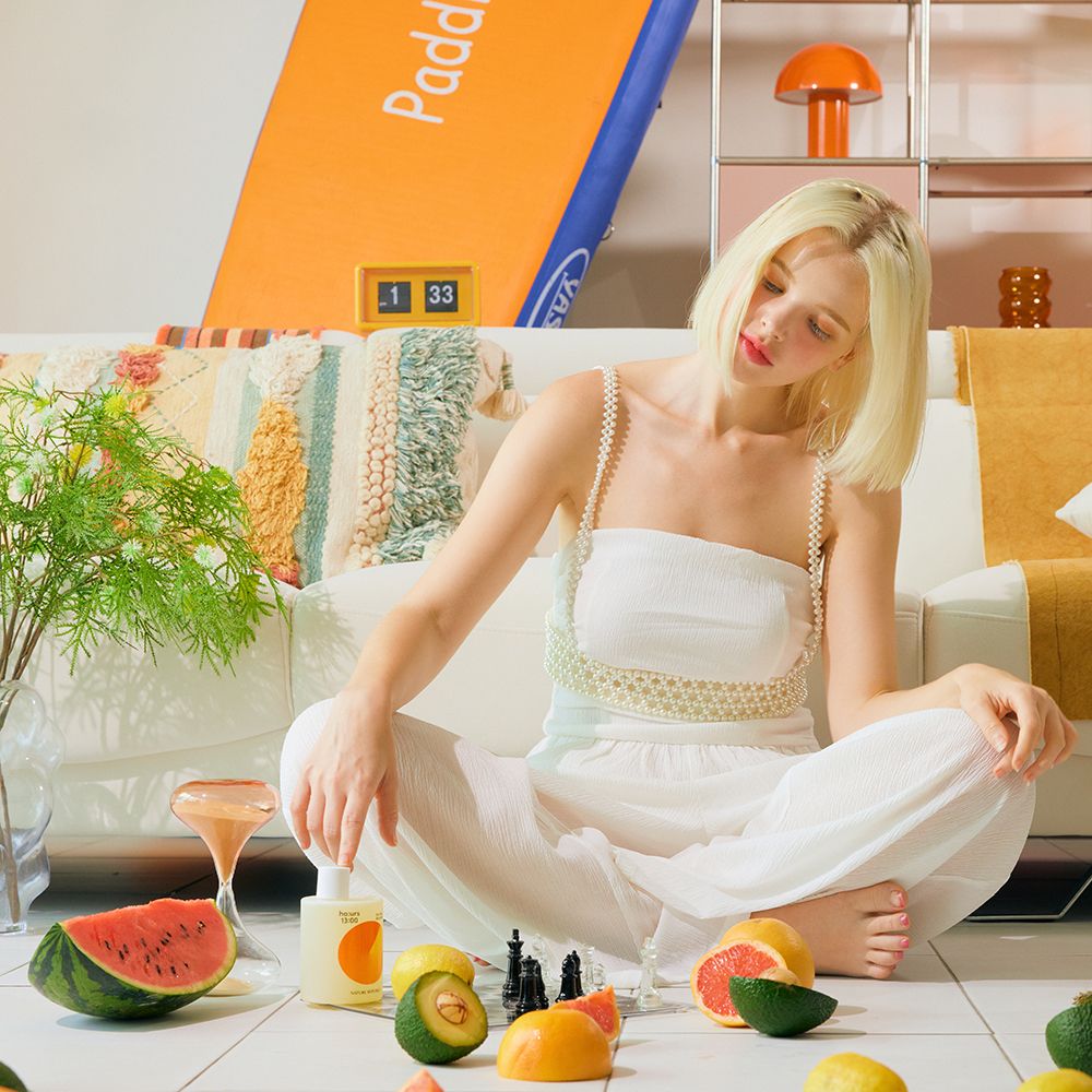 Frau sitzt auf dem Boden. Vor ihr: Obst, Flasche: ho:urs, Sun Step Body Wash. Hintergrund: Sofa, Dekorationen.