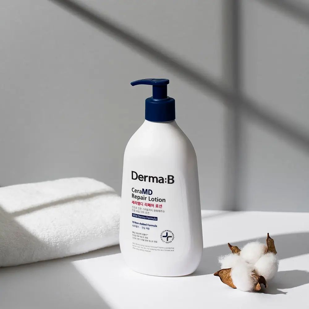 Weiße Flasche mit blauem Pumpkopf. Aufschrift: Derma:B CeraMD Repair Lotion. Neben einem Handtuch und Baumwollblüten.