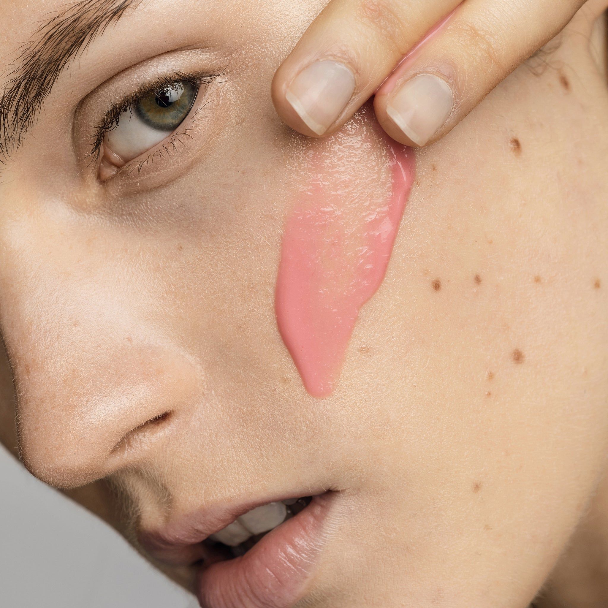 Nahaufnahme eines Gesichts. Rosa Serum wird auf die Wange aufgetragen. Haut mit Sommersprossen.