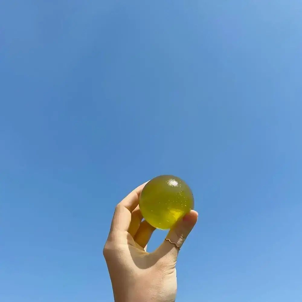 Grünliche Kugel in Hand gehalten, vor blauem Himmel. Hand mit Ring. Klare, runde Form.