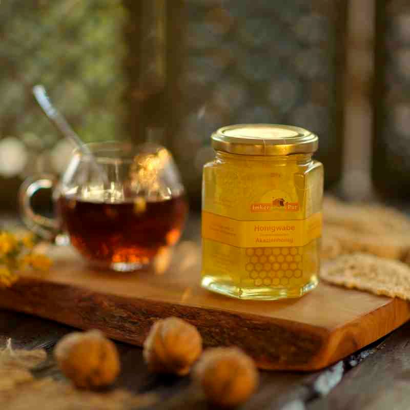 Ein Glas Honigwabe von ImkerPur, neben einer Tasse Tee und Walnüssen. Das Glas ist sechseckig und enthält Honig mit einer Honigwabe. Auf dem Etikett steht "Honigwabe Akazienhonig".