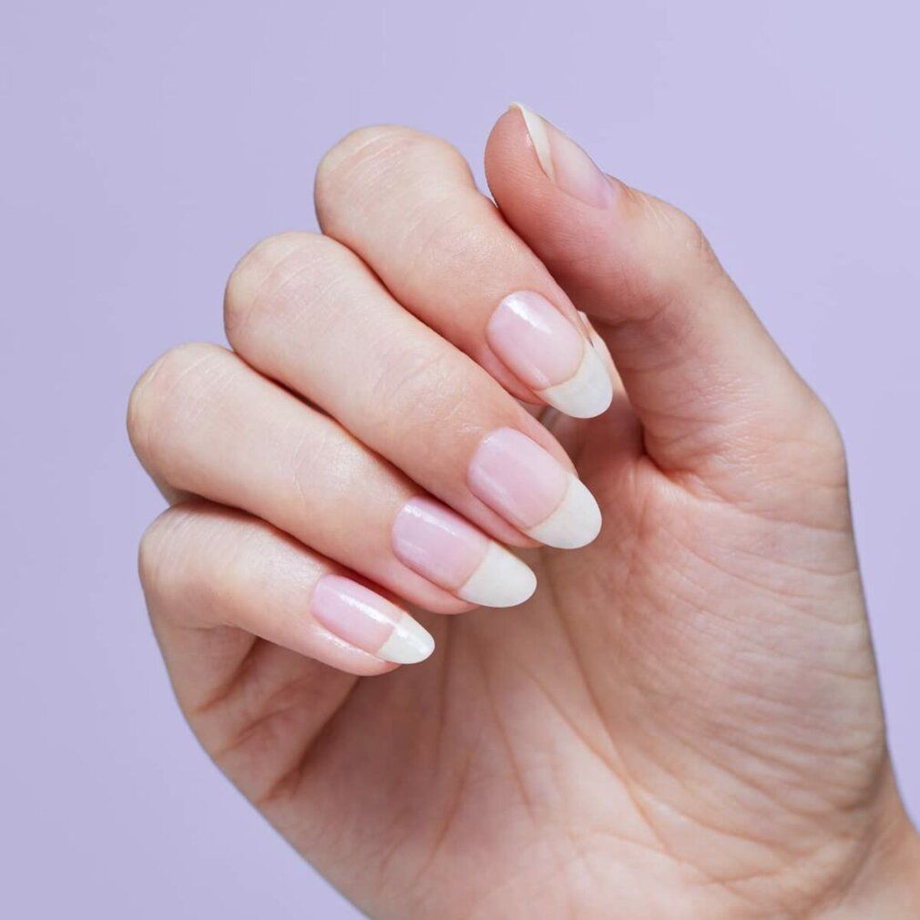 Hand mit French Nails. Die Nägel sind oval und haben eine rosa Basis mit weißen Spitzen. Der Hintergrund ist lila.
