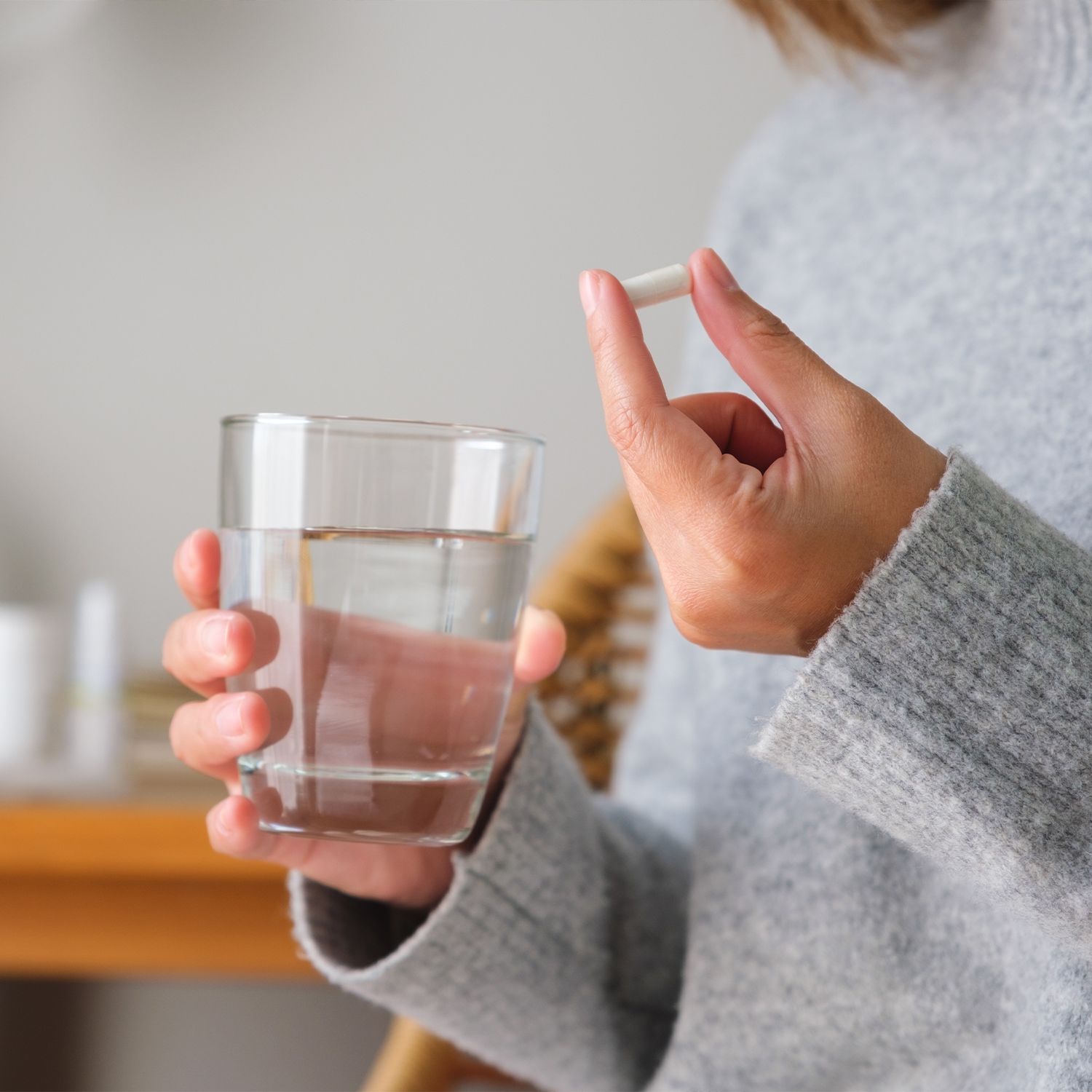 Person hält eine weiße Kapsel über einem Glas Wasser. Person trägt einen grauen Pullover. Helle Umgebung.