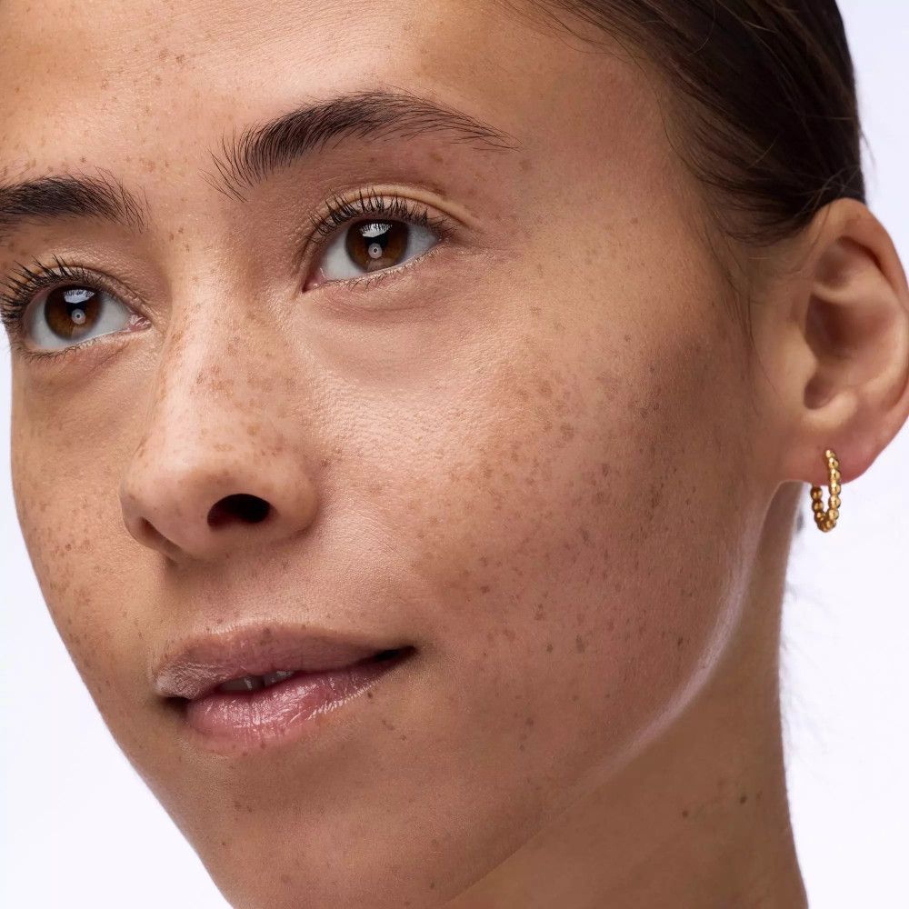 Porträt einer Frau mit Sommersprossen. Sie trägt goldene Ohrringe.