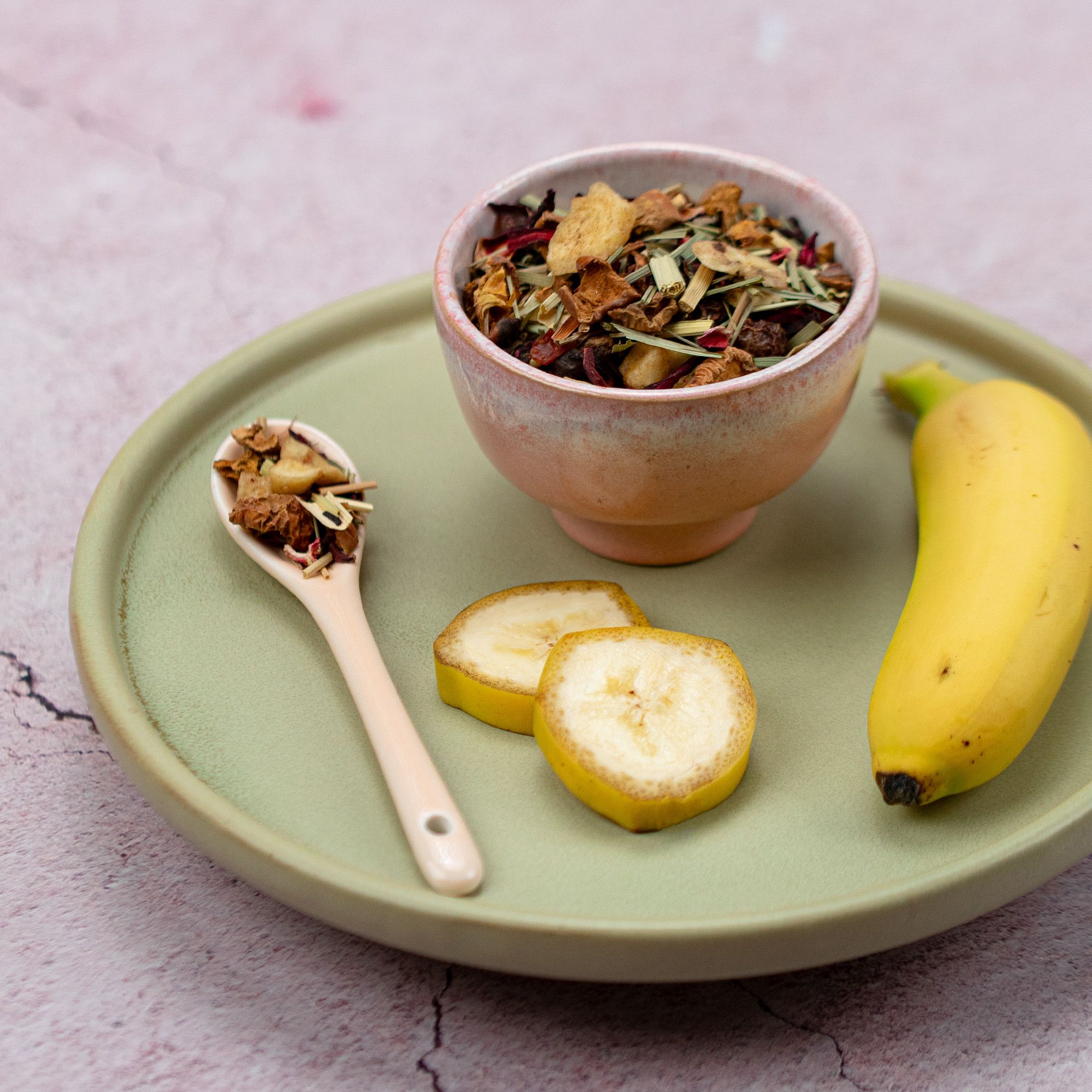 Schale und Löffel mit losem Tee, Bananenscheiben, ganze Banane auf Teller. Rosa und grüne Farbtöne.