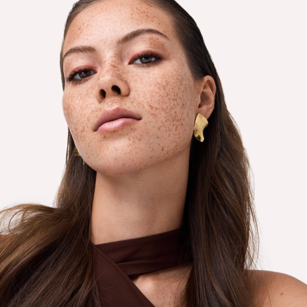 Porträt einer Frau mit Sommersprossen. Sie trägt goldene Ohrringe und rotes Augen-Make-up.